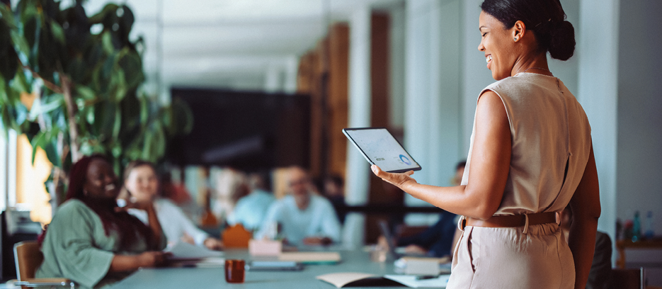 Confident businesswoman presenting to colleagues in a modern office setting. She is holding a digital tablet showing graphs and data, while colleagues are engaged and attentive.