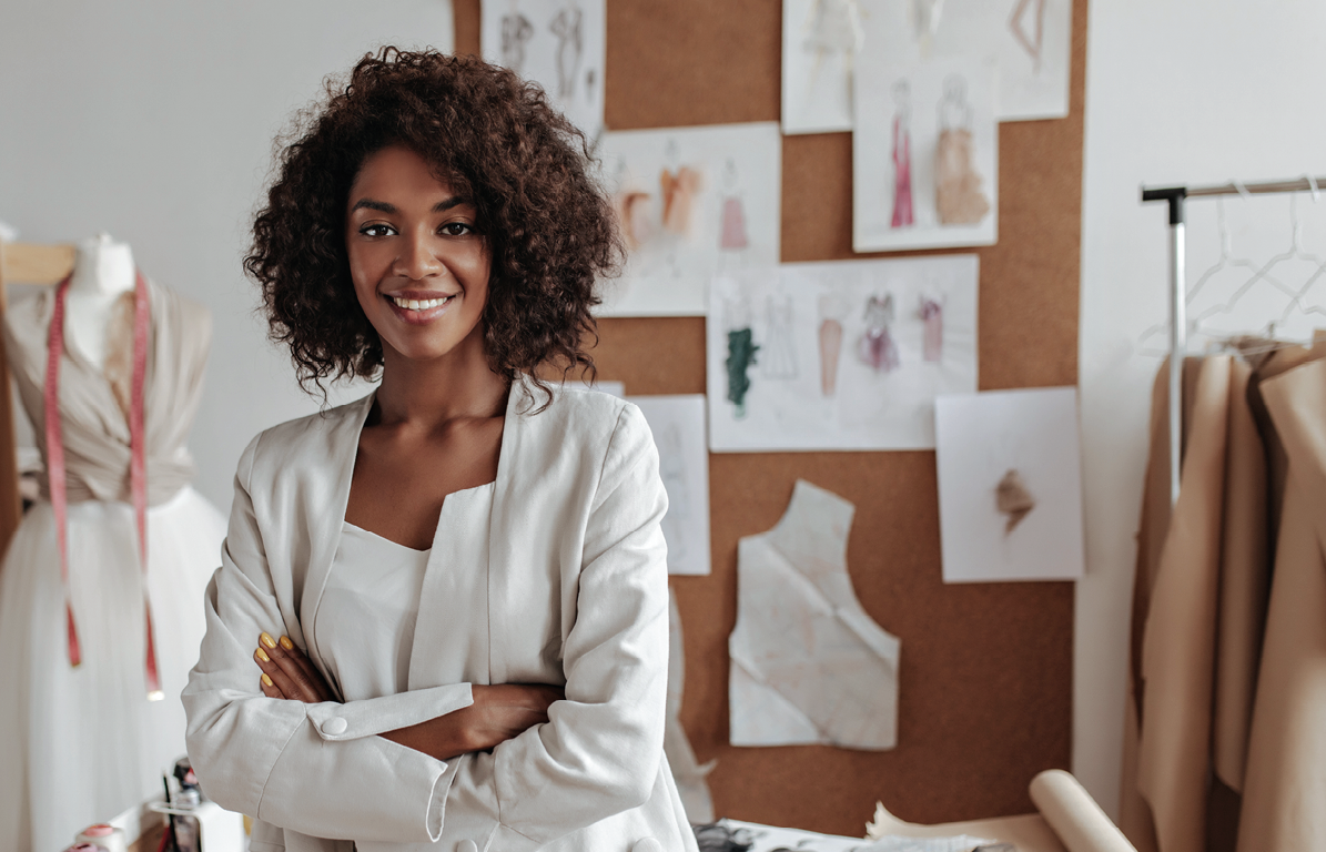 Beautiful curly brunette dark-skinned fashion designer poses in office, leans on table. Young lady in white suit crosses arms and smiles.