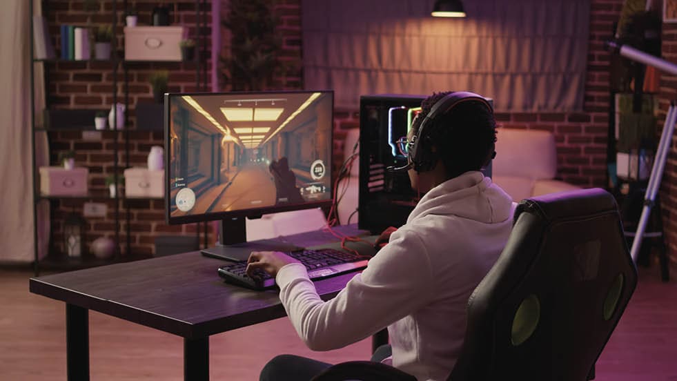 Over shoulder view of african american gamer using pc gaming setup relaxing playing first person shooter in multiplayer. Man streaming while explaining gameplay in online action game talking to team.